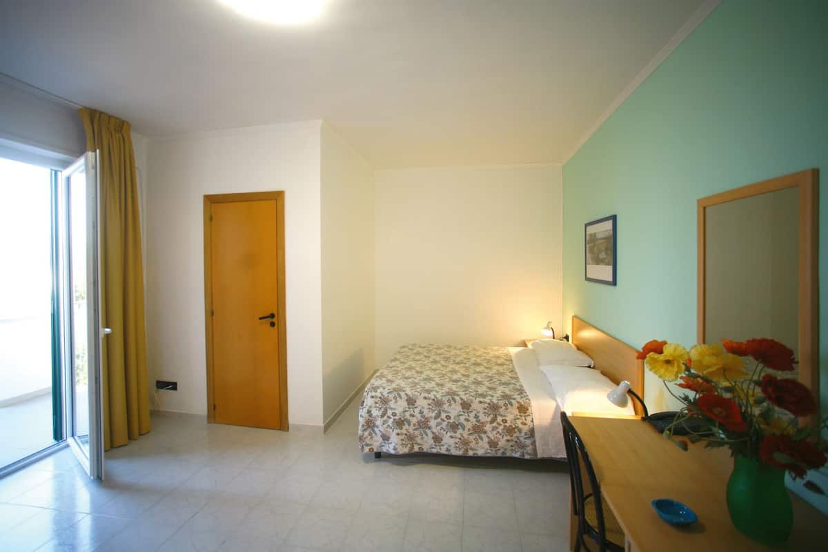 A bedroom with a bed, a desk with a vase of flowers, a door, and a window with curtains.