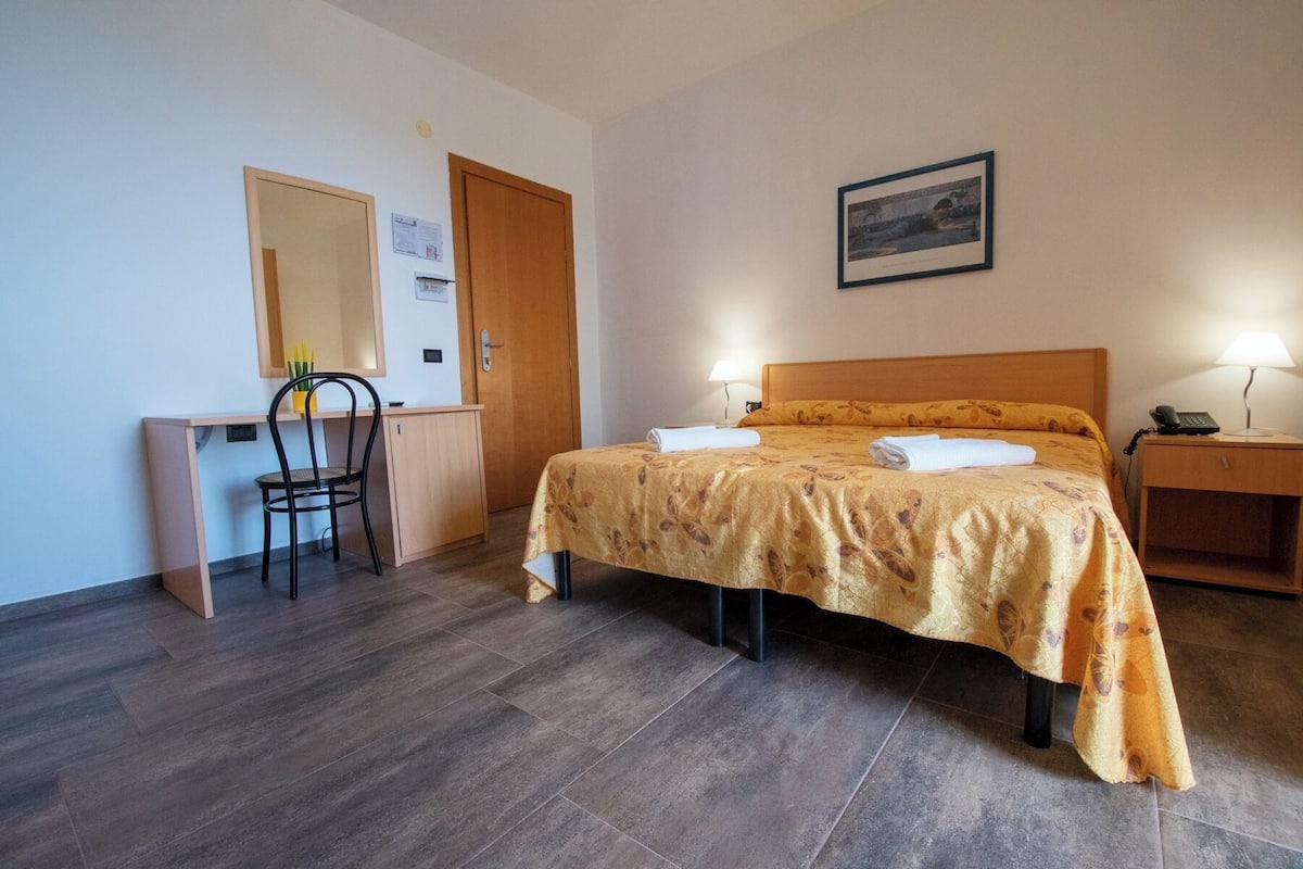 A hotel room with a bed, a desk, a chair, a nightstand, and a lamp.