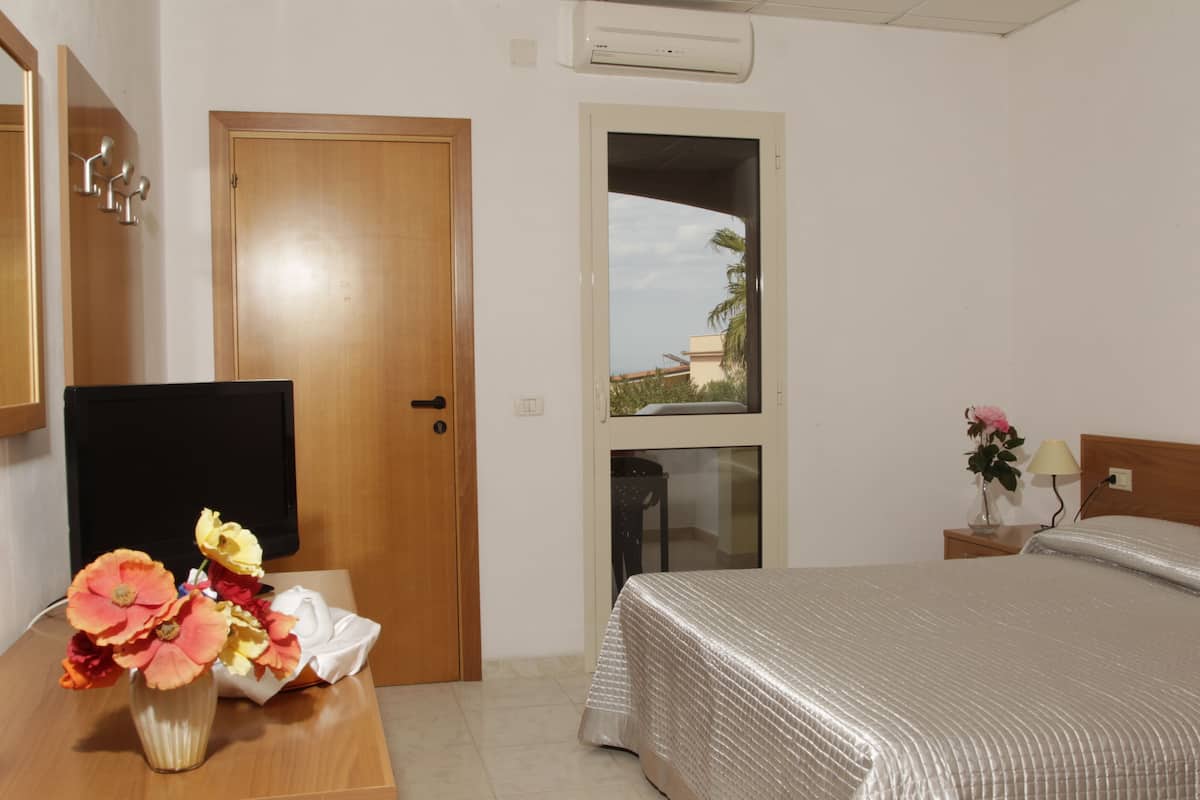 A hotel room with a bed, a wooden door, a TV, a vase with flowers, and a window with a view of buildings and palm trees.
