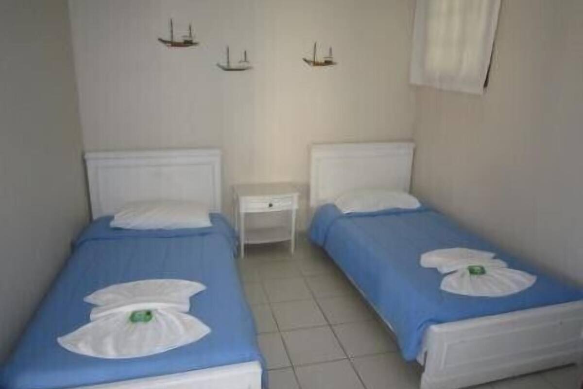 A small, clean room with two beds, each with a blue bedspread and white pillows. There are two bedside tables and a small nightstand.