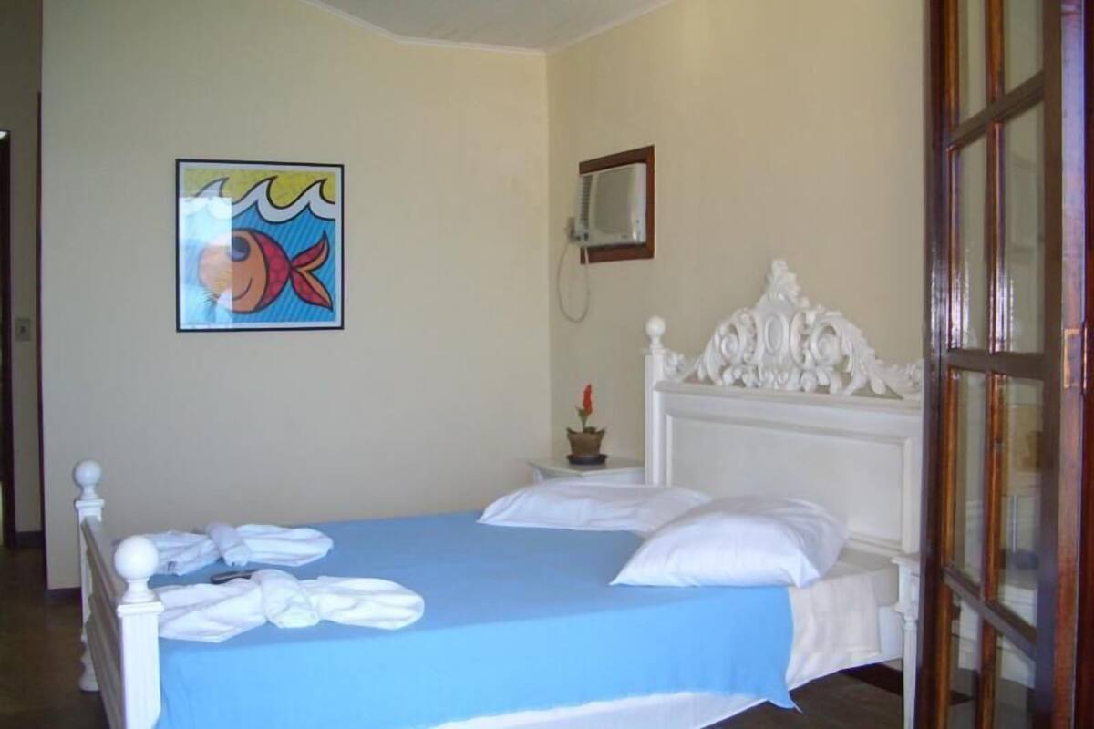 A hotel room with a bed, a blue bedspread, a white headboard, a painting on the wall, and a small table with a flower vase.