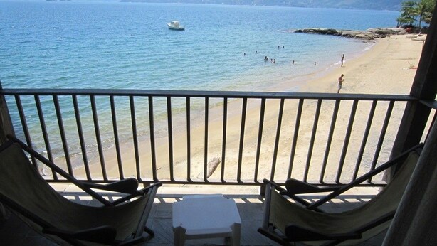 A balcony with two folding chairs and a small table, overlooking a beach with people and a boat in the water. - Pousada Pier 7400 (Angra dos Reis)