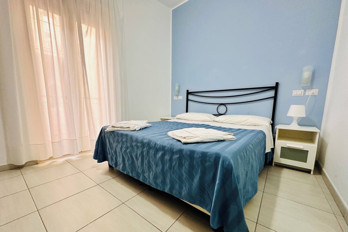 Una cama bien hecha con colcha azul y almohadas blancas, una mesita de noche blanca con lámpara, y una ventana con cortinas de gasa.