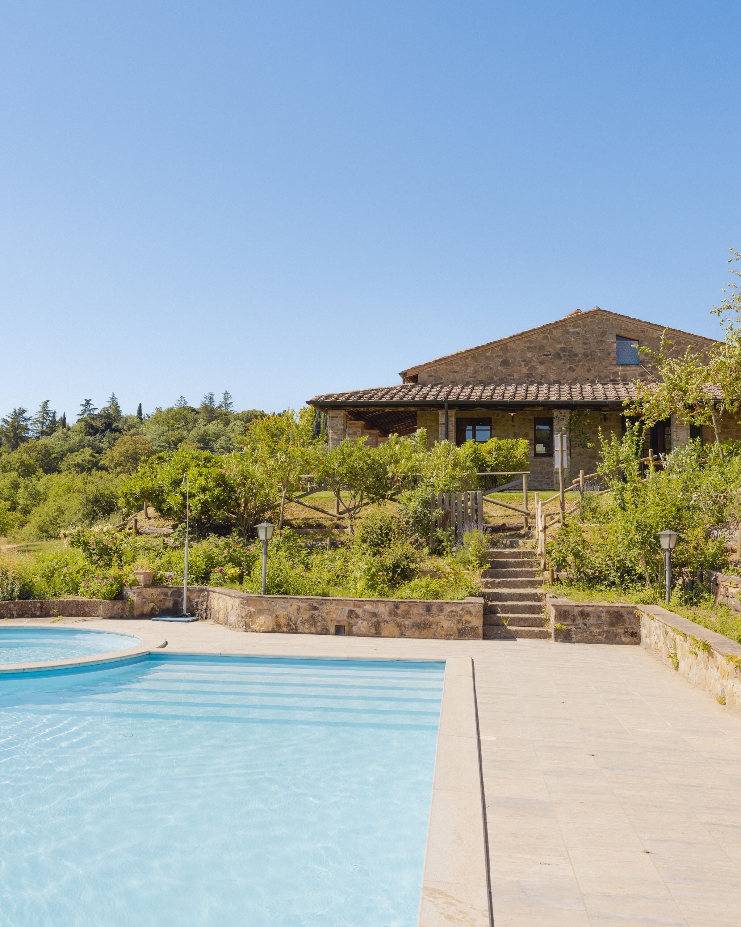 A stone house with a swimming pool, surrounded by greenery and a clear sky.