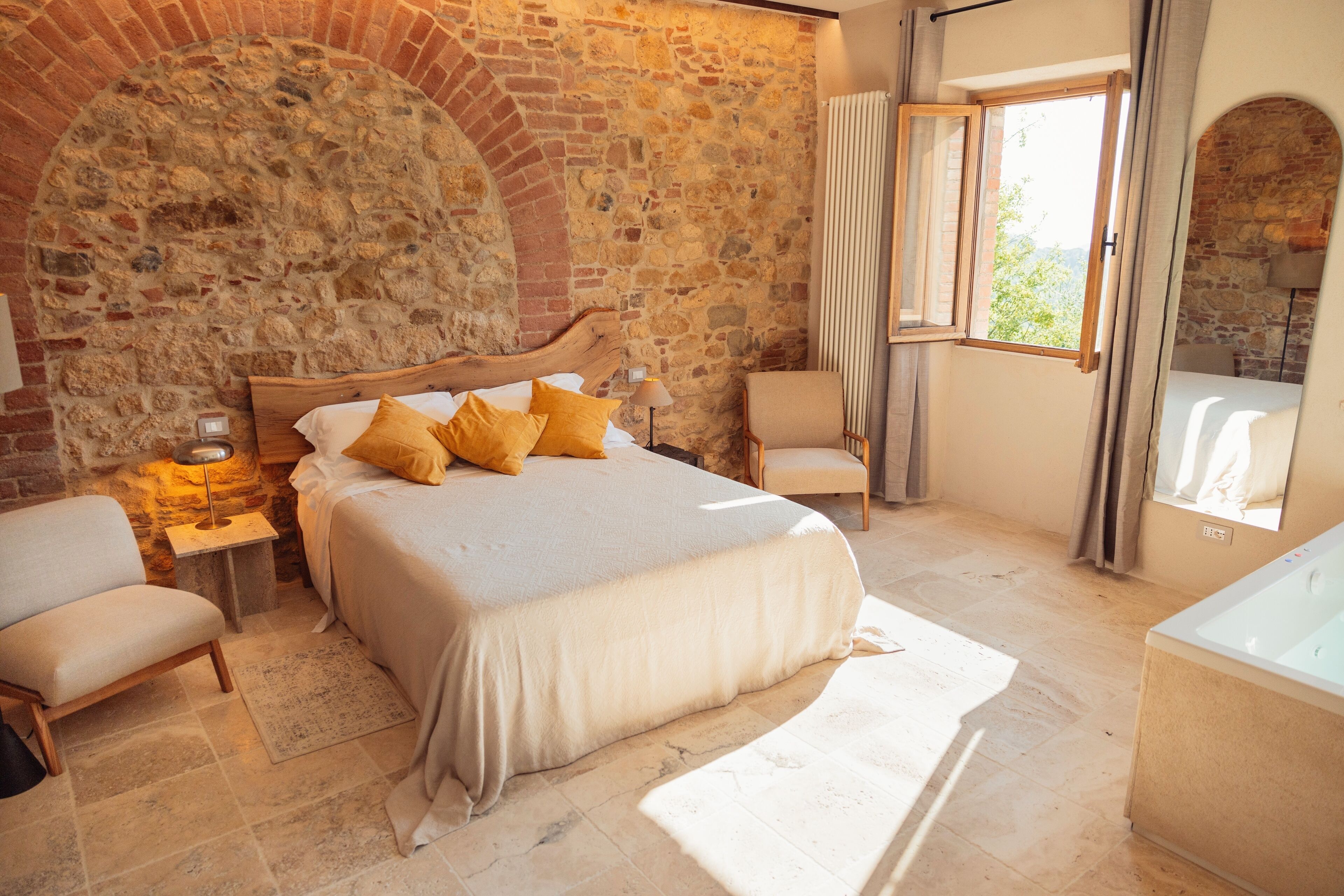 Un dormitorio con pared de piedra, cama de madera, una silla, mesita de noche y ventana.