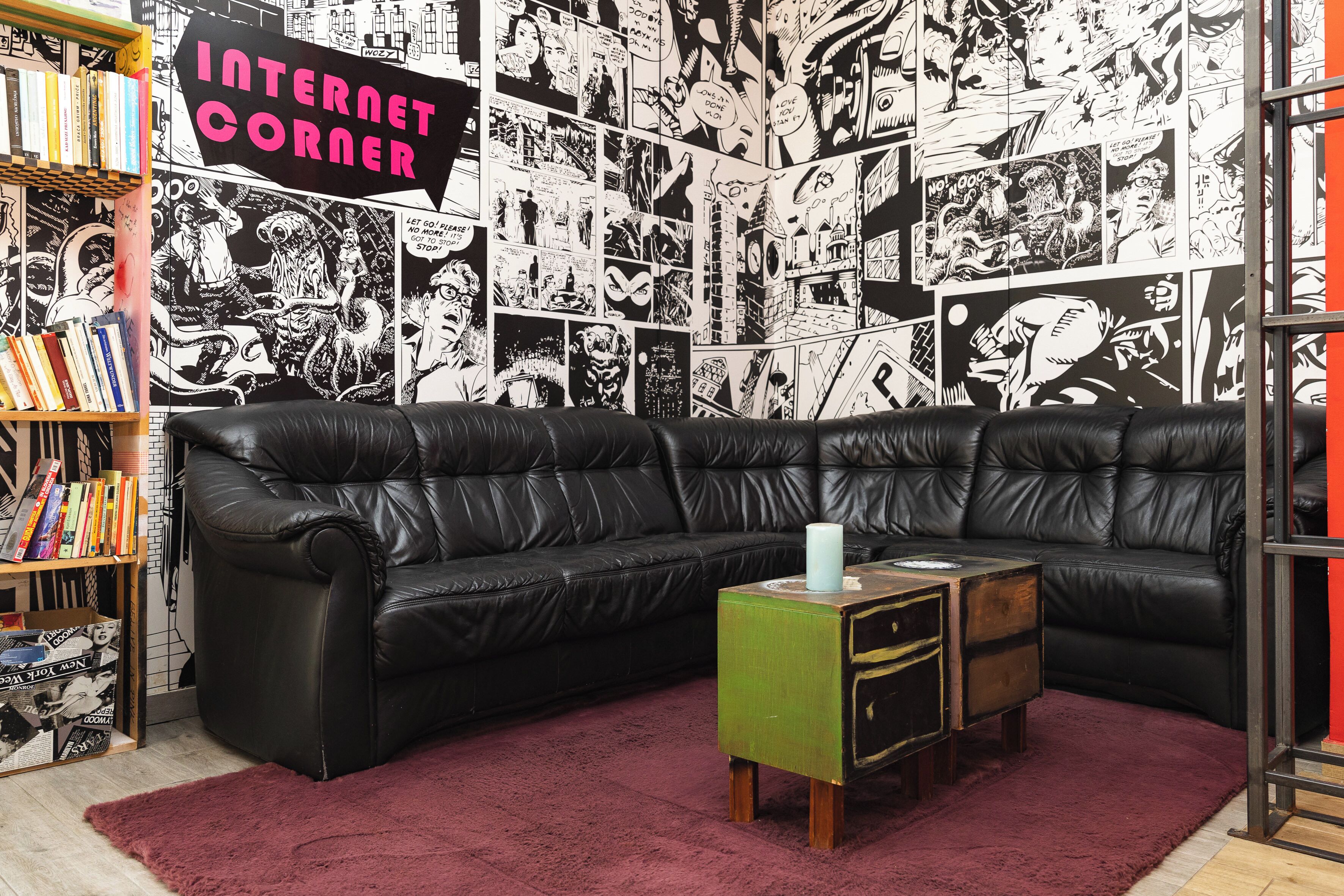 A room with a black leather sectional sofa, a green side table, and a red rug. There is a bookshelf with various books and a sign that reads "INTERNET CORNER".