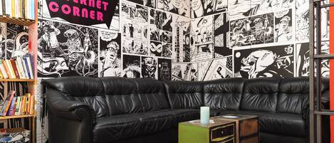 A room with a black leather sectional sofa, a green side table, and a red rug. There is a bookshelf with various books and a sign that reads "INTERNET CORNER".