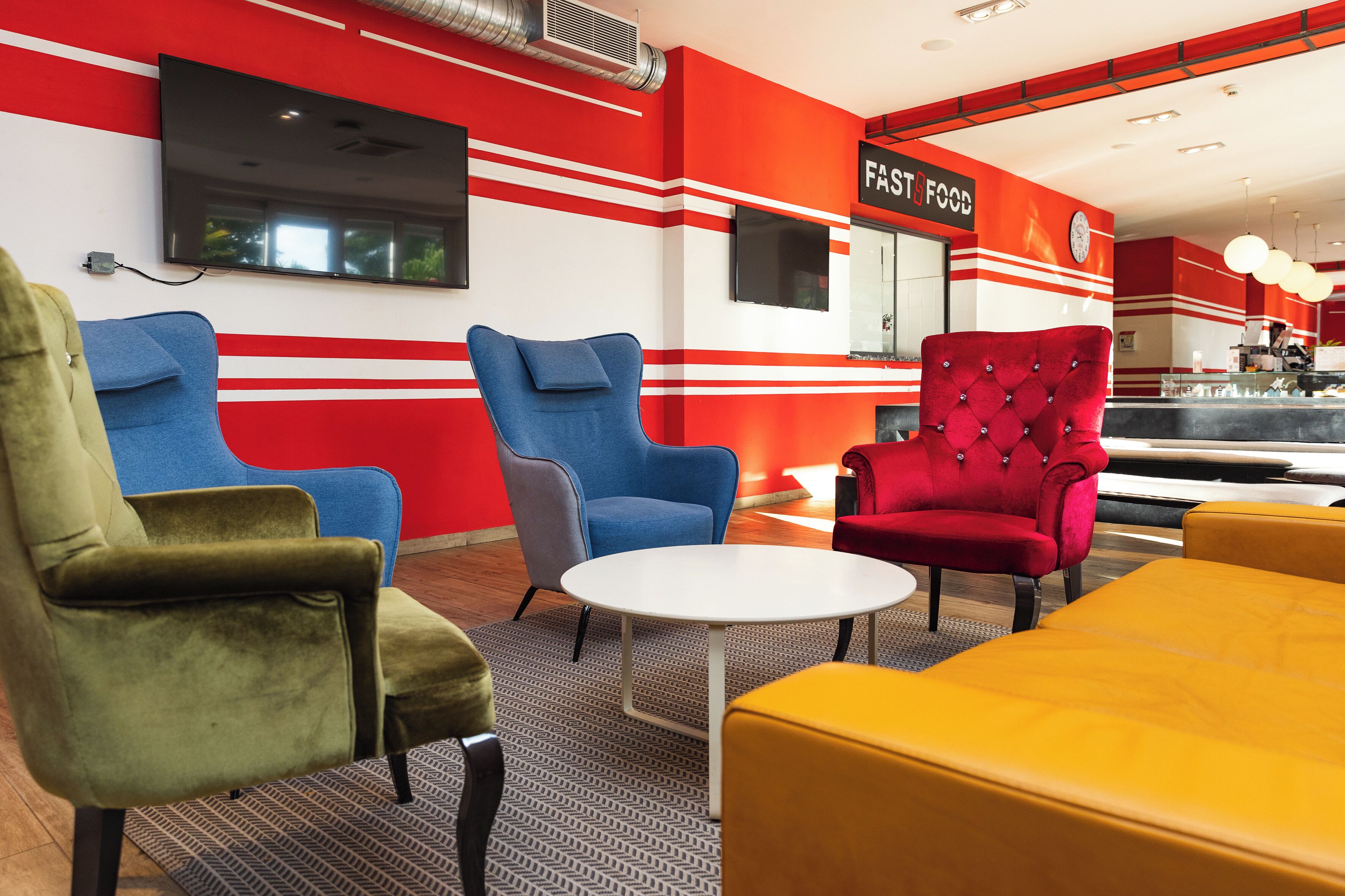A modern interior with red and white striped walls, a flat-screen TV, and a 'FAST FOOD' sign. There are three upholstered chairs and a yellow ottoman.