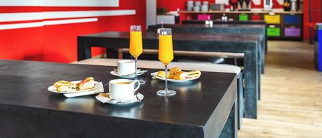 Zona de dinar d'un hostal modern, amb taules, aliments per l'esmorzar i una paret vermella amb el nom de l'hostal.