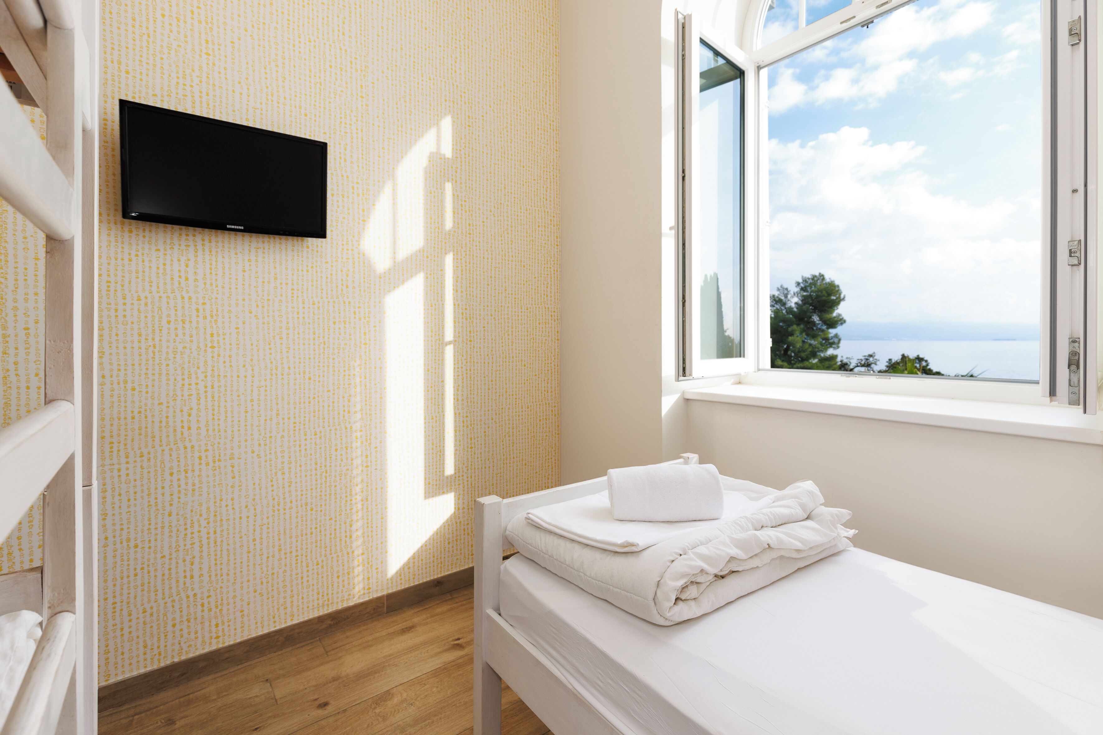 A hotel room with a bed, a flat-screen TV mounted on the wall, and a window with a view of the sea.