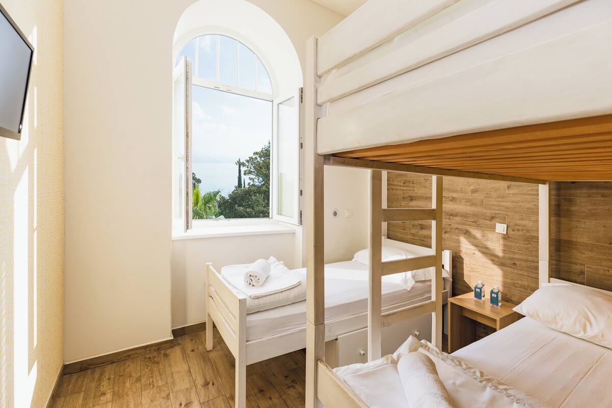 A bunk bed room with a wooden headboard, a small bedside table with a lamp, and a window with a view of greenery.