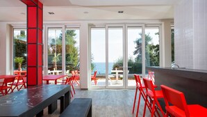 A modern dining area with red chairs and tables, large glass doors leading to a terrace with a view of the sea. - Hostel Link  (Lovran)
