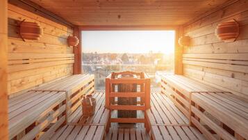 Un sauna con bancos de madera y una mesa central, ventanas amplias con vista a edificios y paredes de madera.