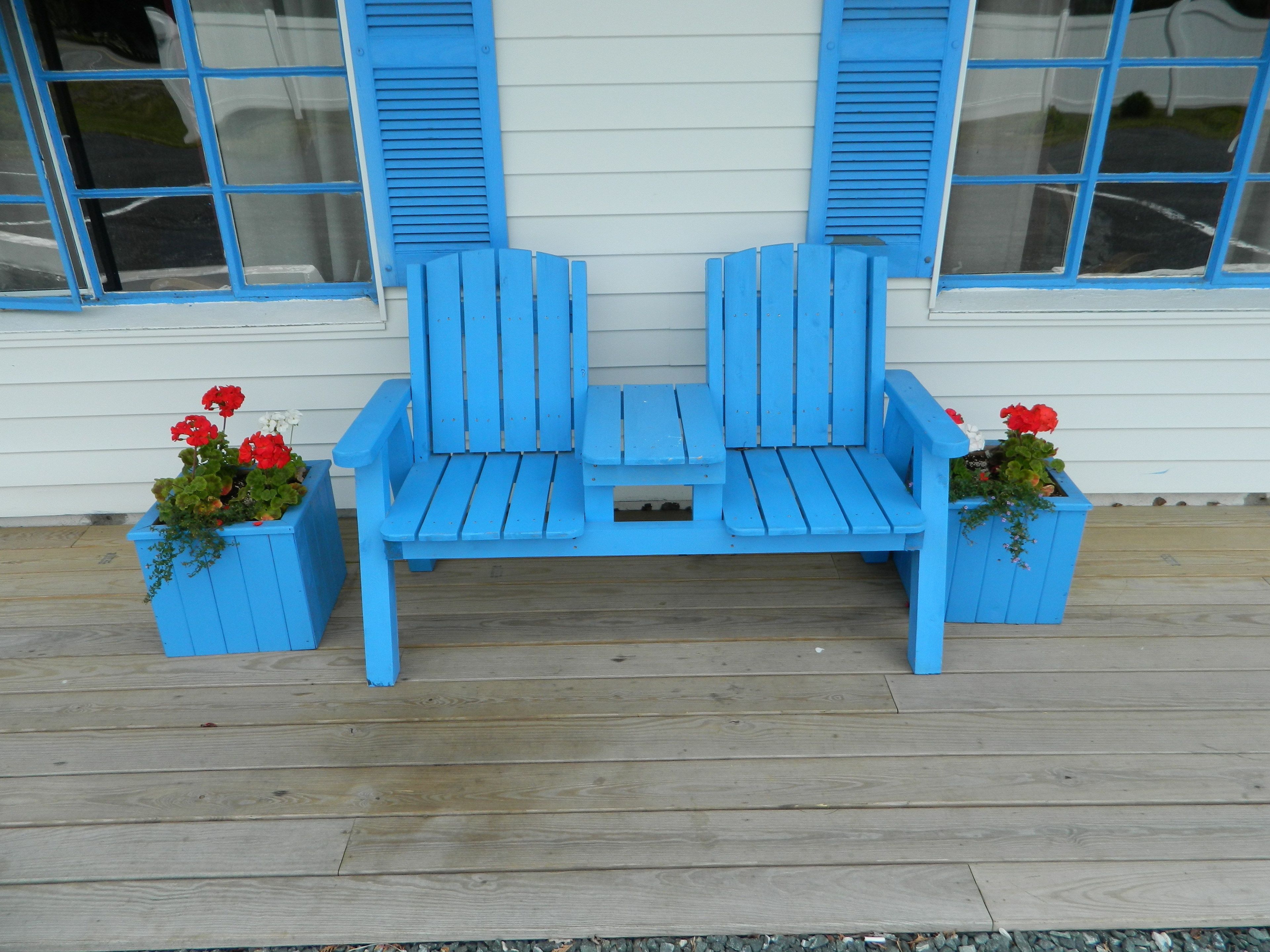 A blue Adirondack chair set on a wooden deck in front of a white building with blue shutters.