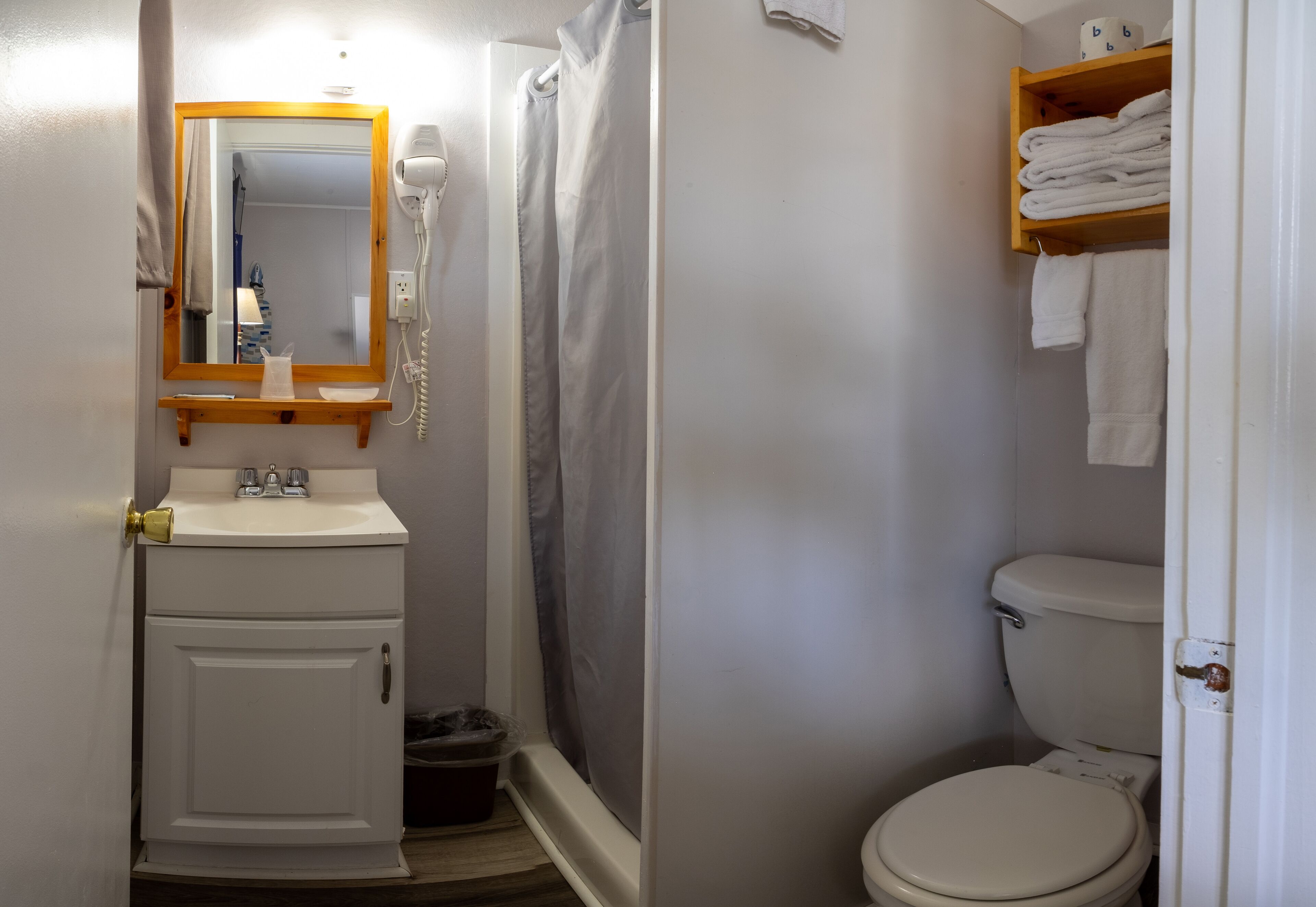 A small bathroom with a white sink, a mirror, a shower curtain, a toilet, and a shelf with folded towels.
