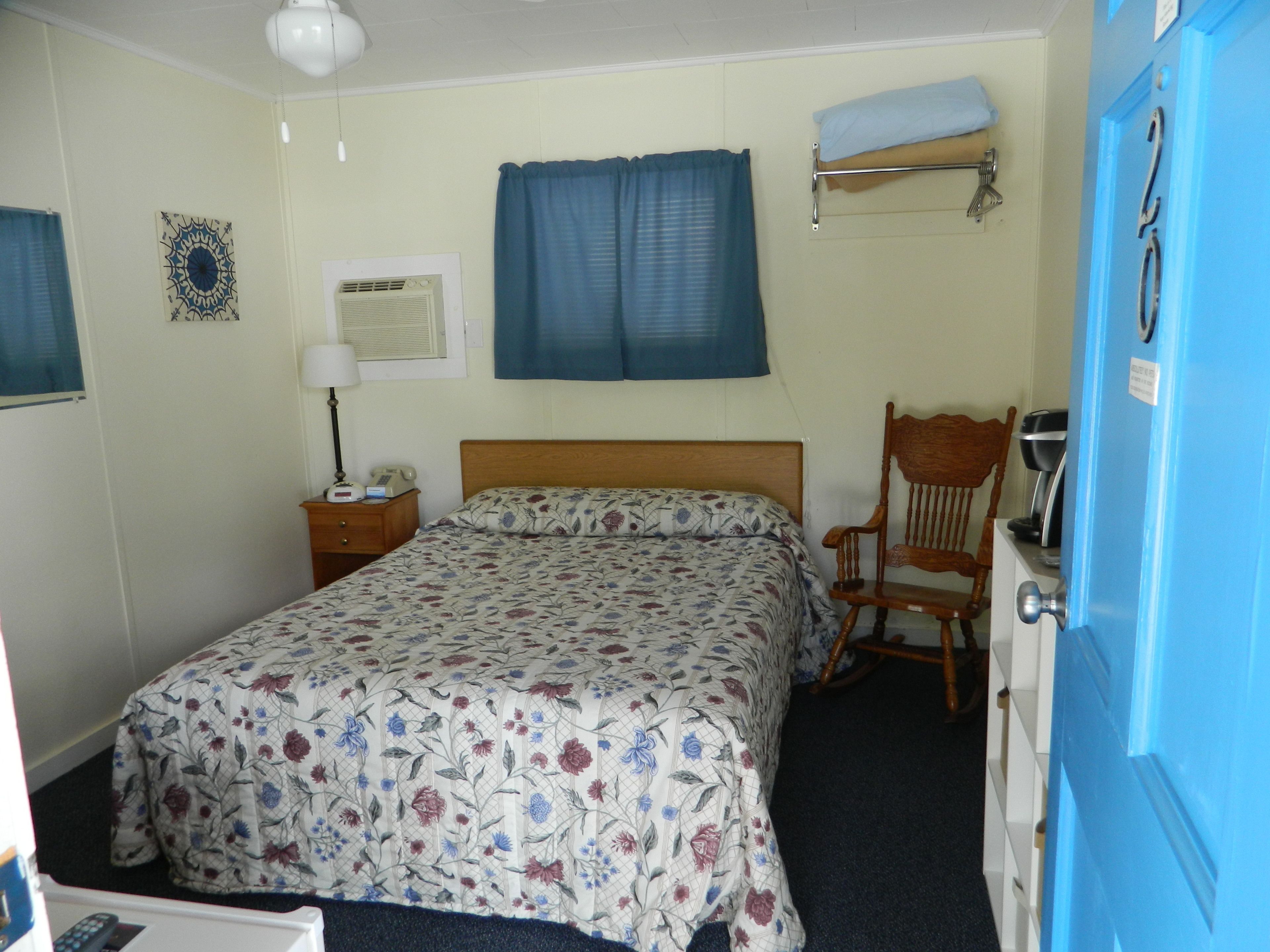 A bedroom with a bed, a chair, a nightstand, a lamp, a wall clock, and a blue door.