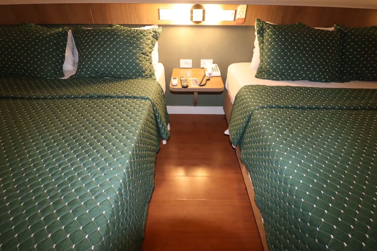 A cabin with two beds, each with green quilted bedding and pillows, a small table with a phone and remote control, and a wooden floor.