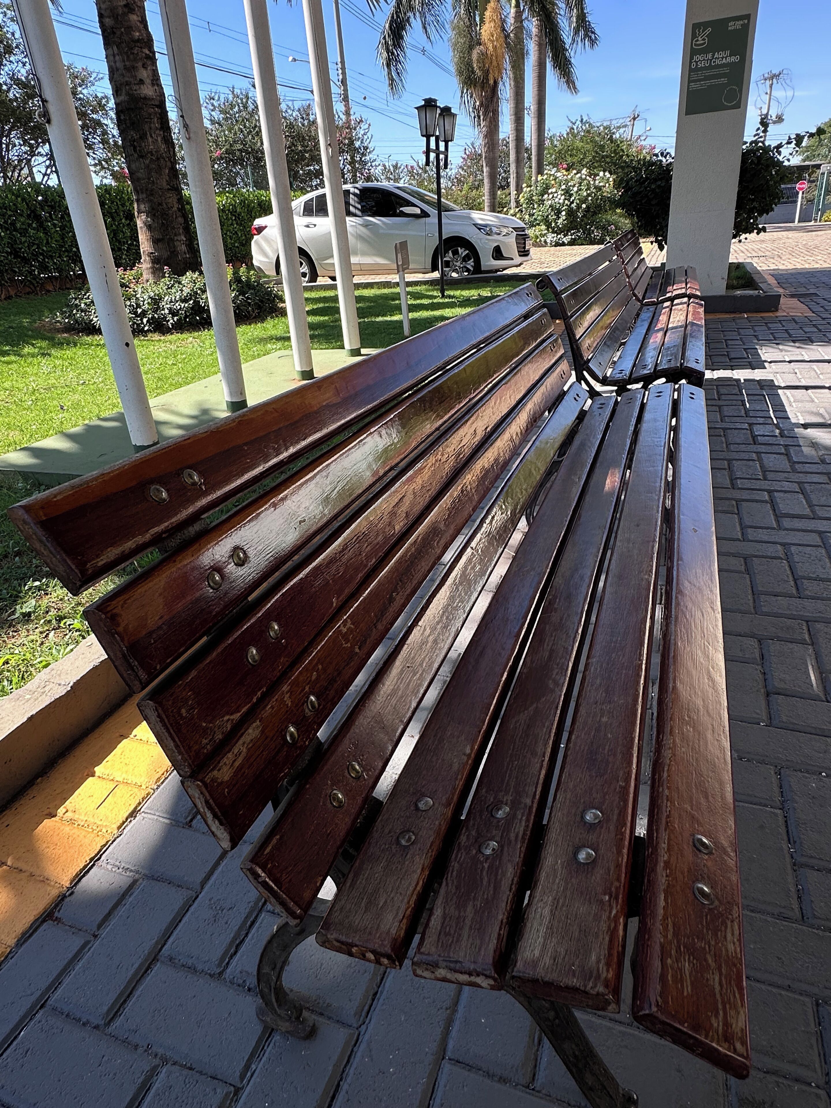 A wooden bench with metal armrests and a metal frame, located on a paved walkway with a grassy area and palm trees in the background.