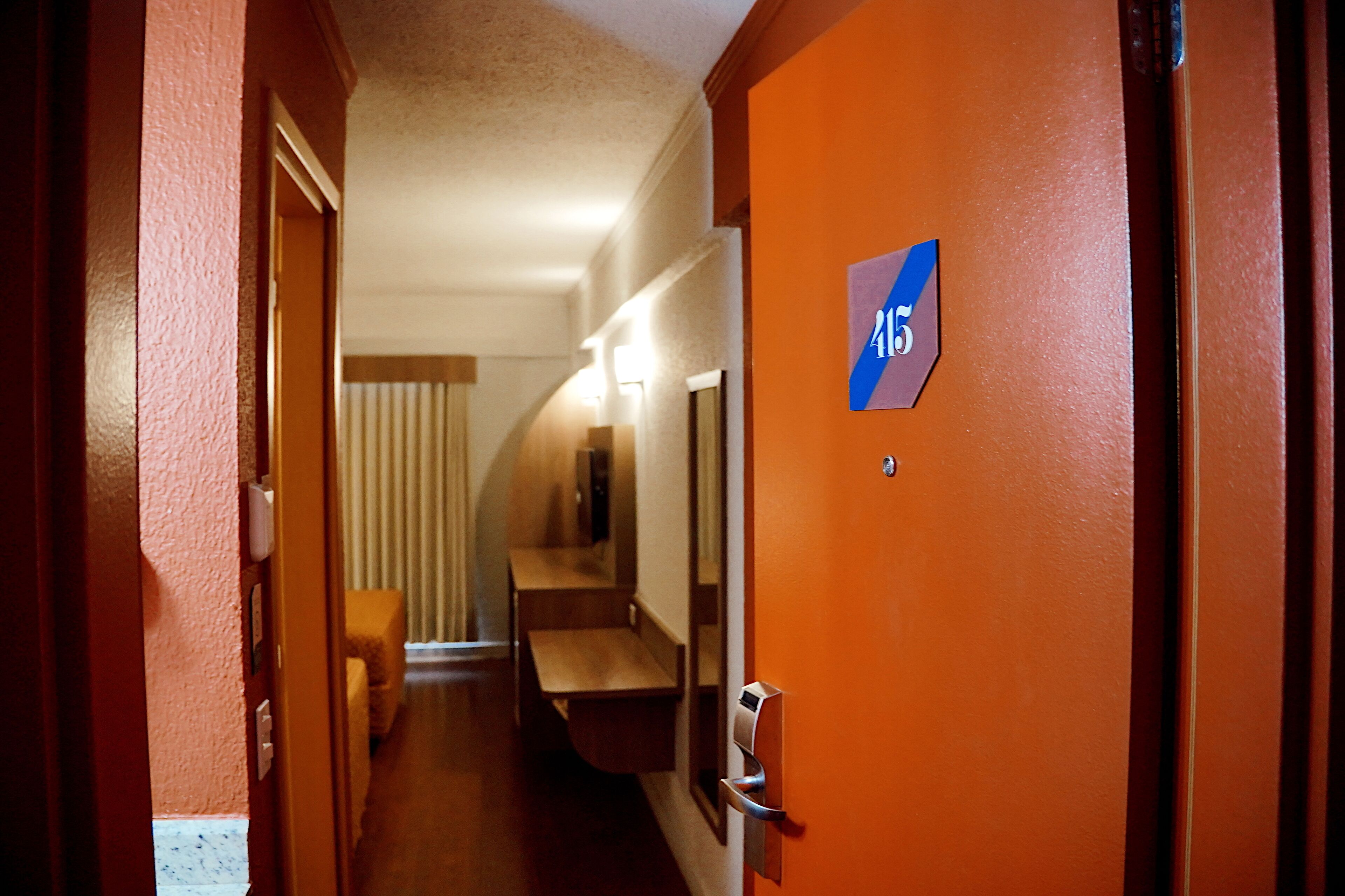 Un pasillo de hotel con un banco de madera, un espejo y una puerta con un letrero azul.