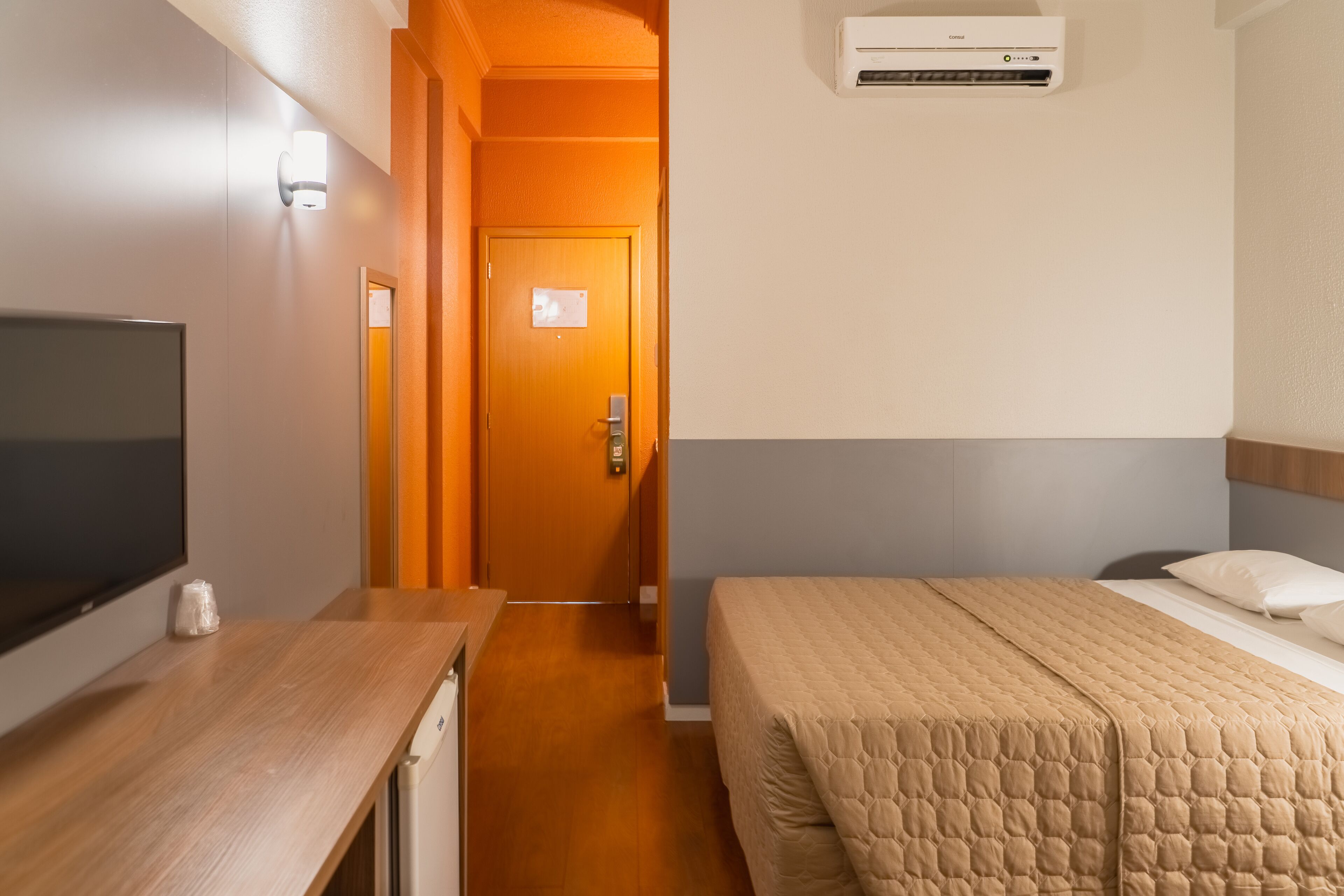Quarto de hotel com cama, televisão, escrivaninha e ar condicionado.