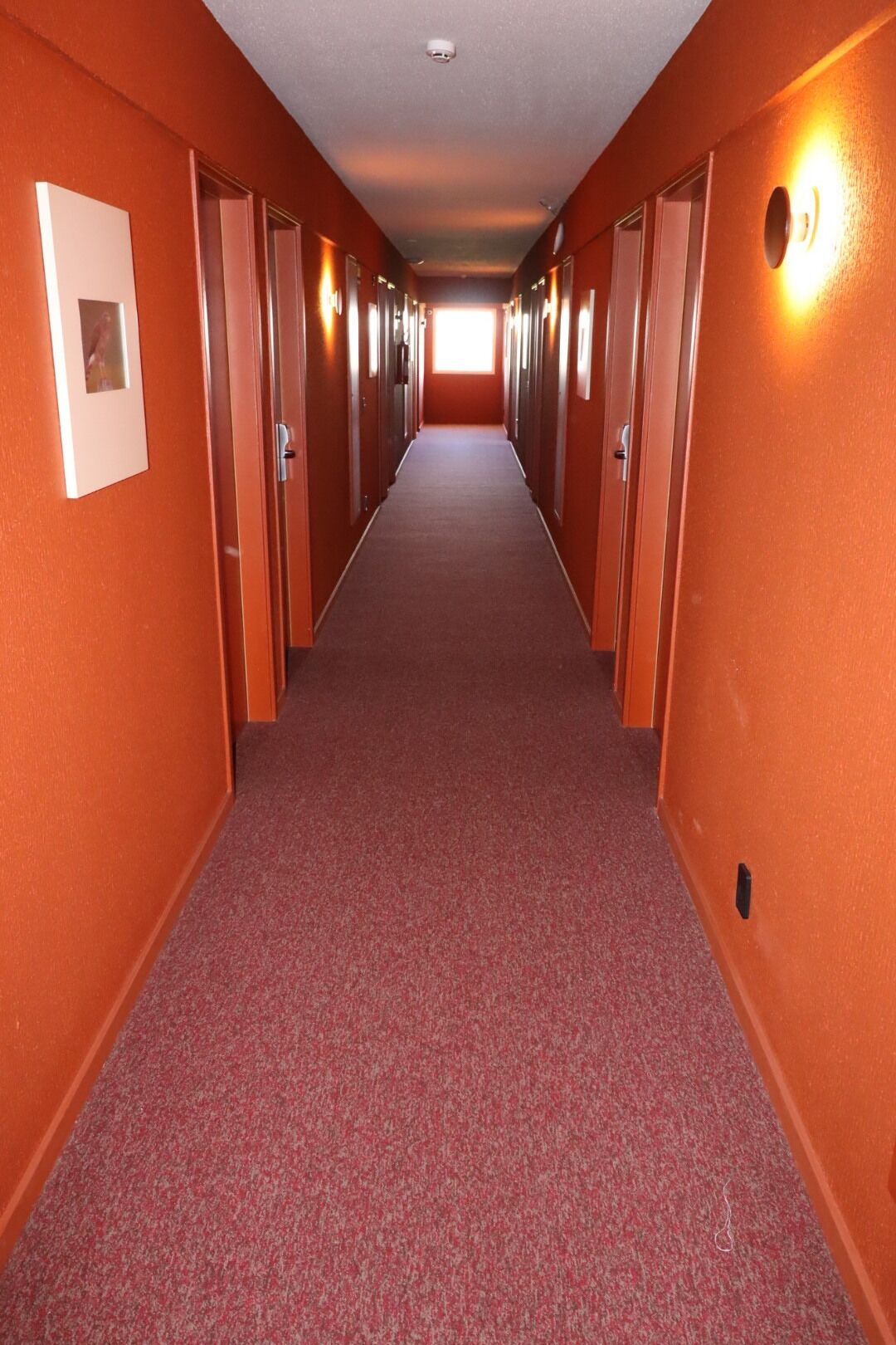 A long, narrow hallway with orange walls and a red carpet, leading to a bright exit at the end.