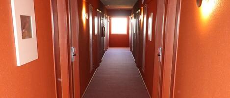 A long, narrow hallway with orange walls and a red carpet, leading to a bright exit at the end.