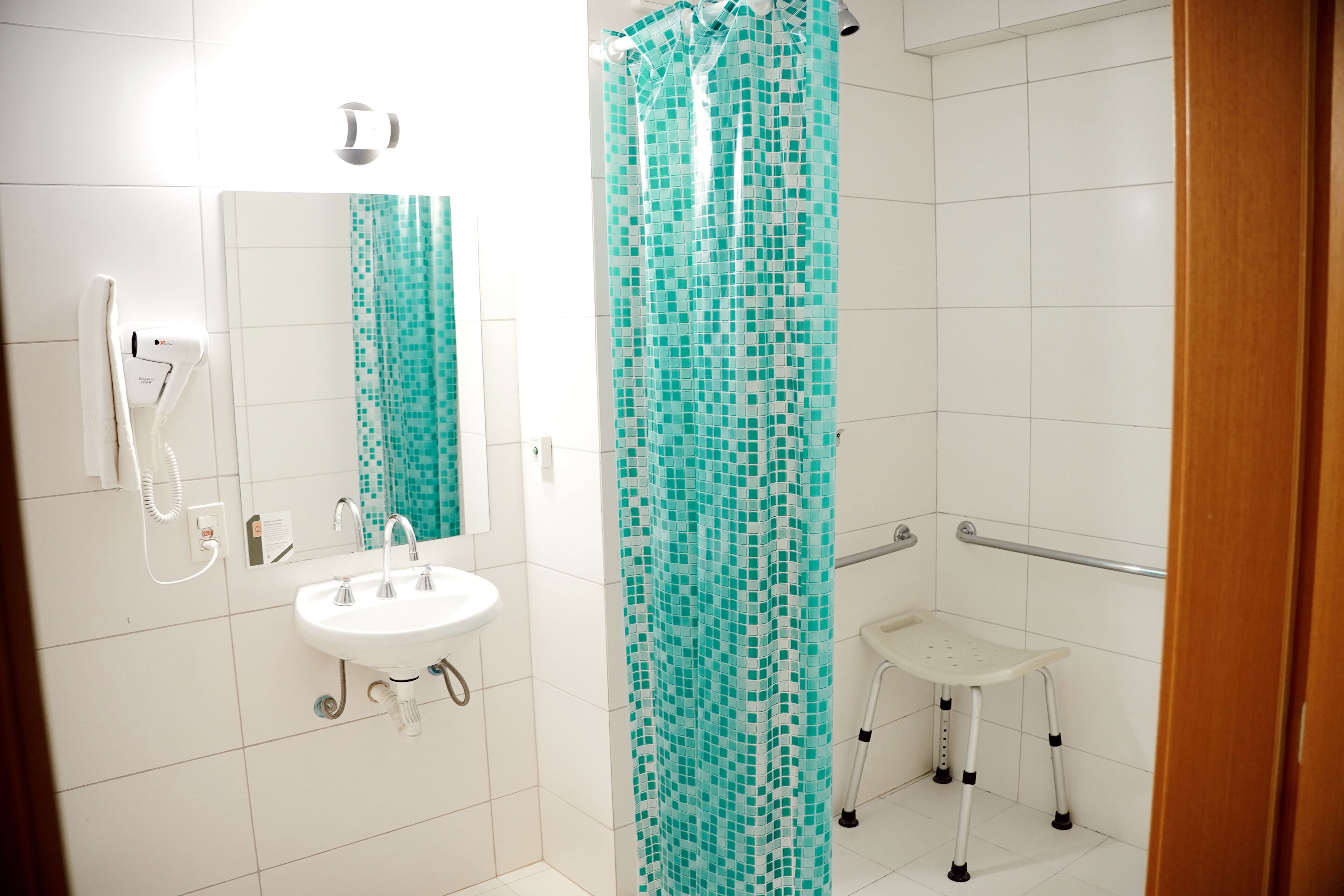 Een badkamer met een wit wastafel, een spiegel, een douche met een turquoise mozaïek gordijn, en een klein wit bankje.