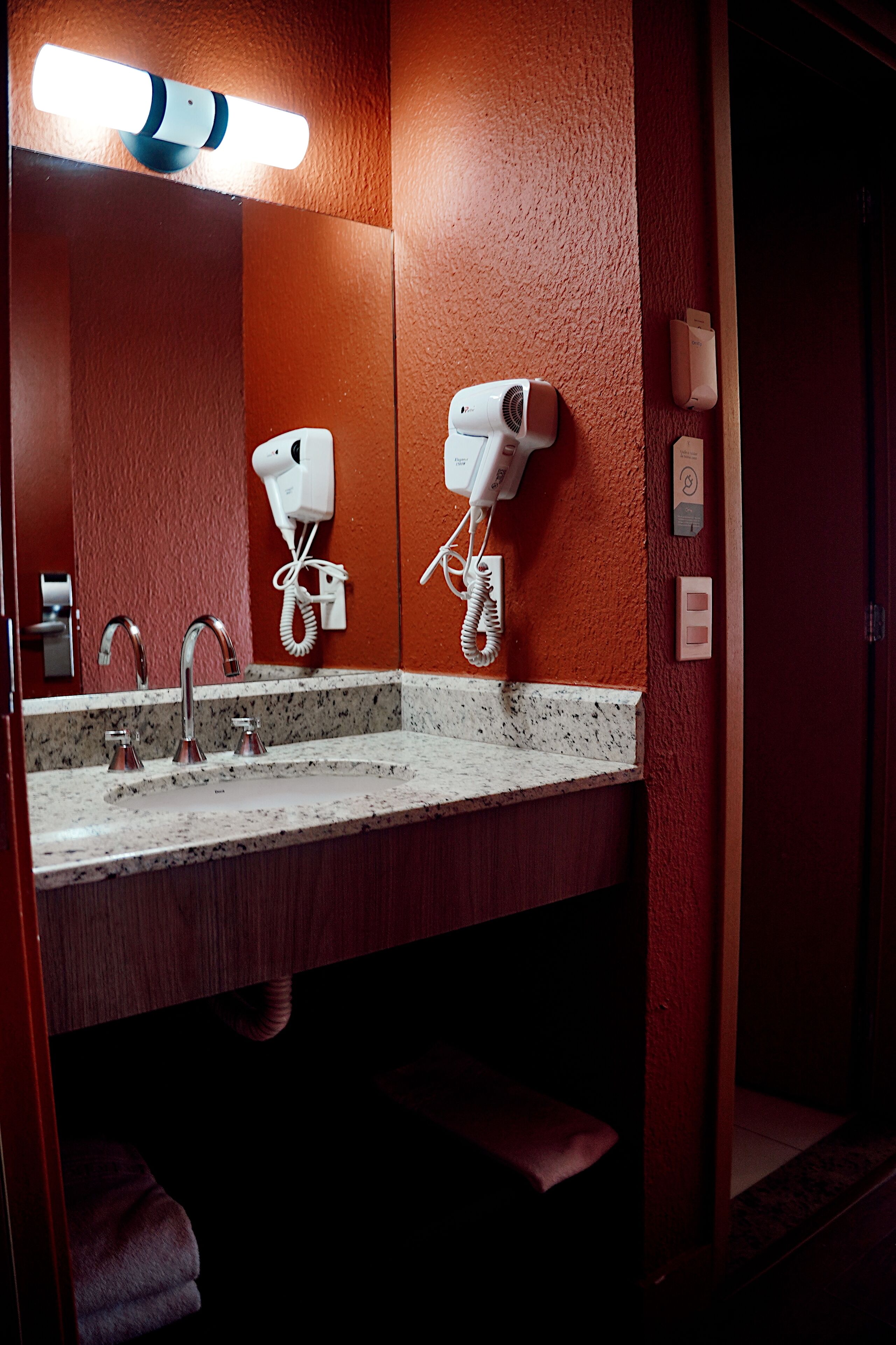 Baño de hotel con lavamanos, dos secadores de pelo y un espejo.