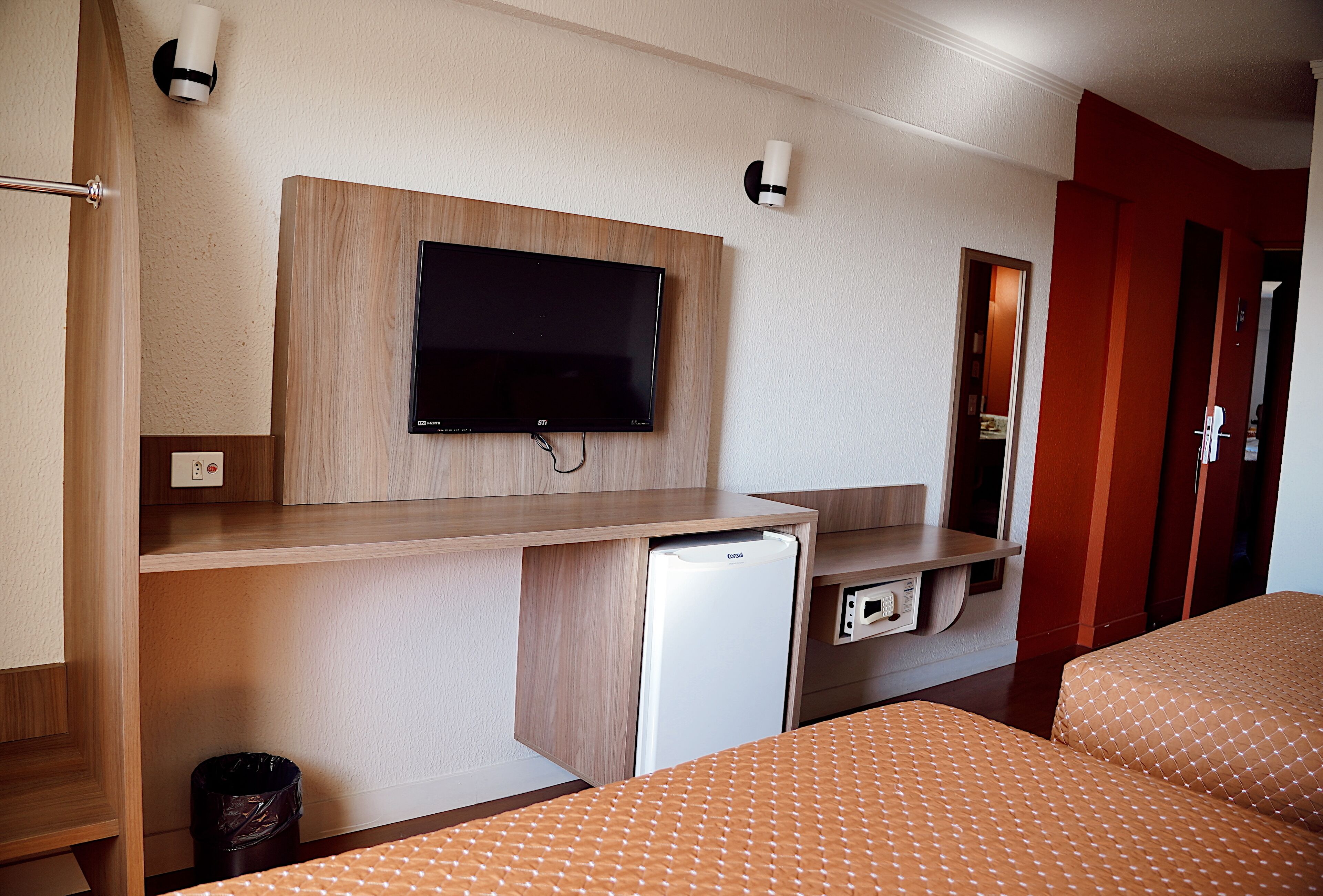 A hotel room with a bed, a TV mounted on a wooden panel, a small refrigerator, and a trash bin.
