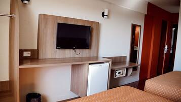 A hotel room with a bed, a TV mounted on a wooden panel, a small refrigerator, and a trash bin.