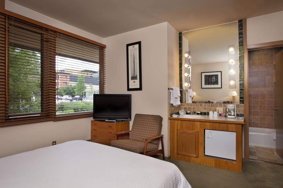 A hotel room with a bed, a TV, a chair, a small desk with a mirror, and a bathroom with a sink and lights.