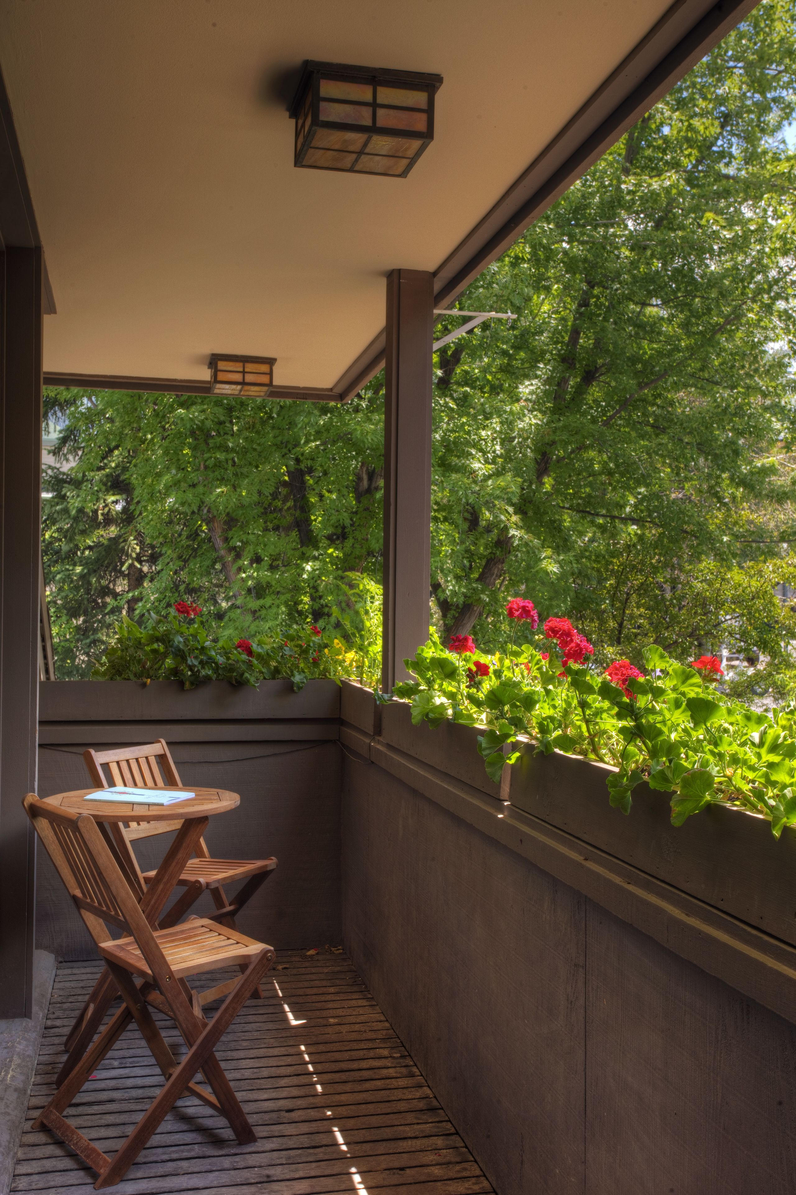 A balcony with wooden chairs, a small table, and a view of greenery and flowers through the window.