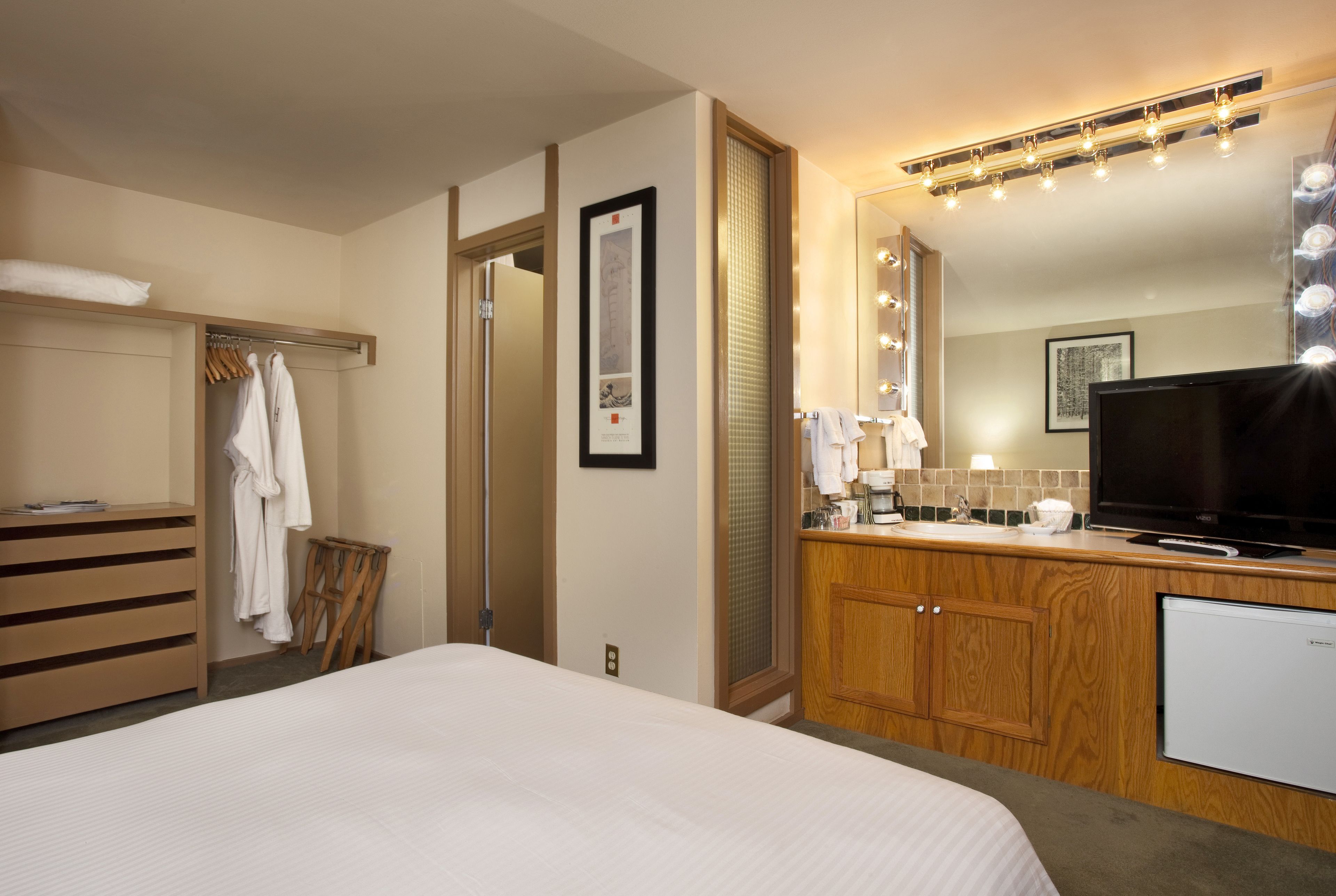 Una habitación de hotel con cama, armario de madera, tocador con espejo e iluminación, televisor y refrigerador pequeño.