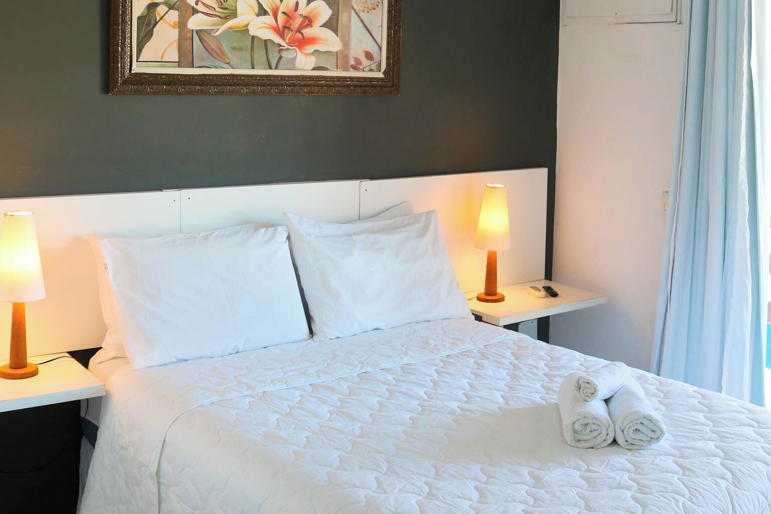 A hotel room with a bed, two bedside lamps, a framed floral artwork, and a window with curtains.