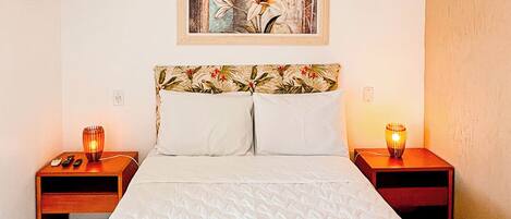 A hotel room with a bed, two bedside tables, a nightstand, a painting on the wall, and an air conditioning unit.