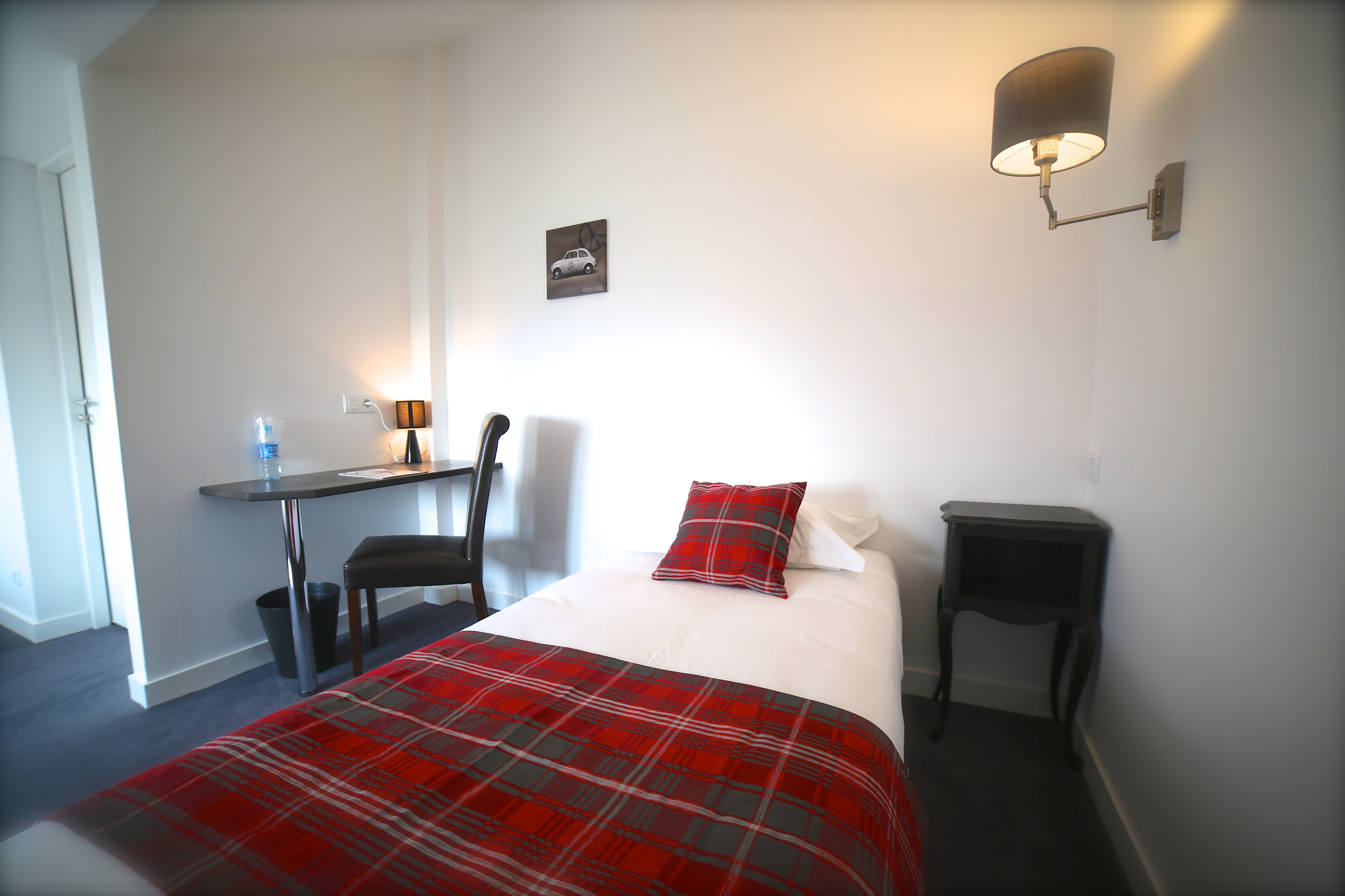 A hotel room with a bed, a desk, a chair, a small table, and a wall-mounted lamp.