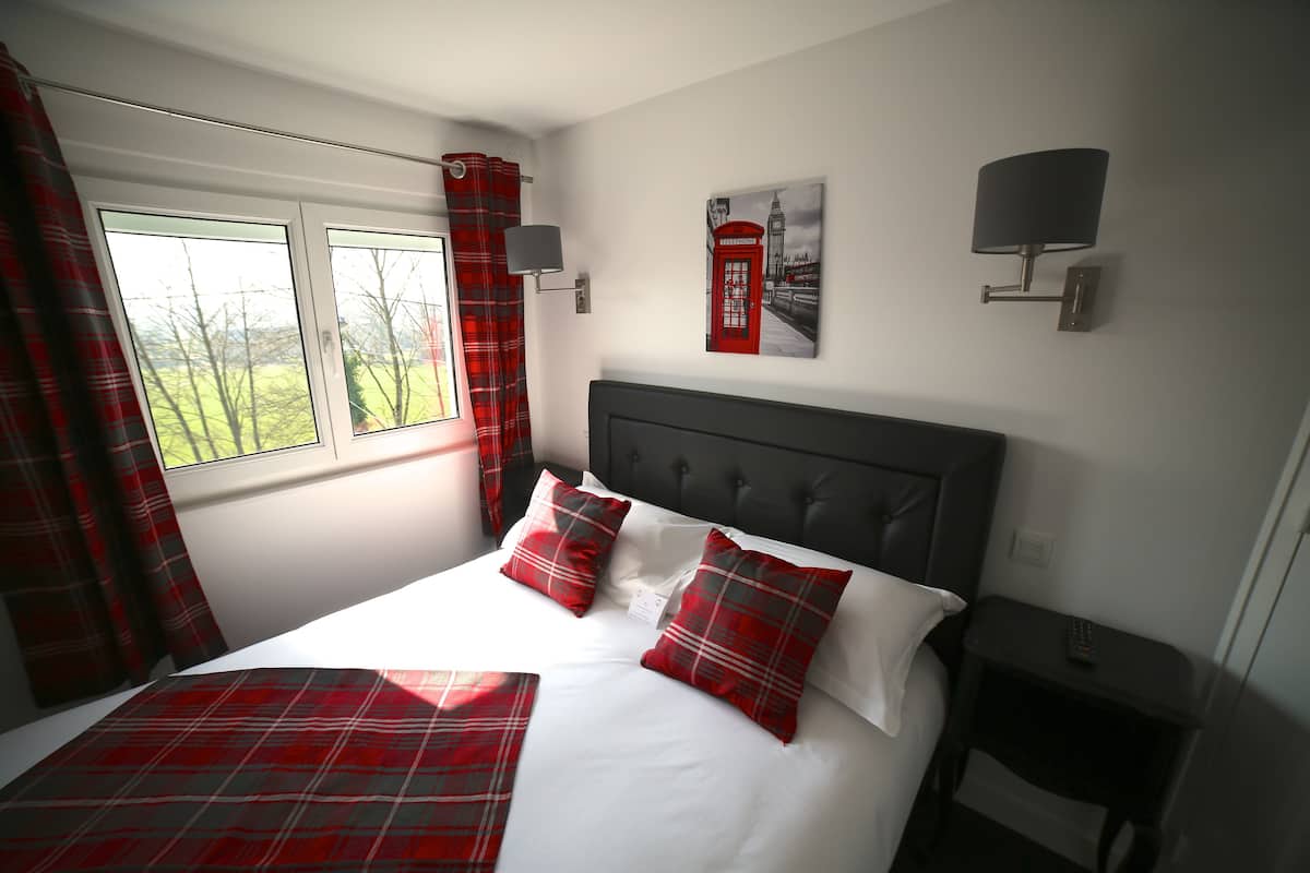 A bedroom with a bed, red and white bedding, a window with curtains, a wall-mounted lamp, and a picture of a red telephone booth.