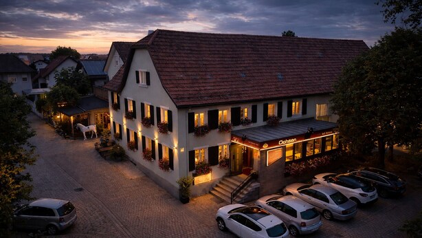 Ein zweistöckiges Gebäude mit rotem Ziegeldach, einem Restaurant namens "Ochsens" im Erdgeschoss und einem Parkplatz mit mehreren Autos.