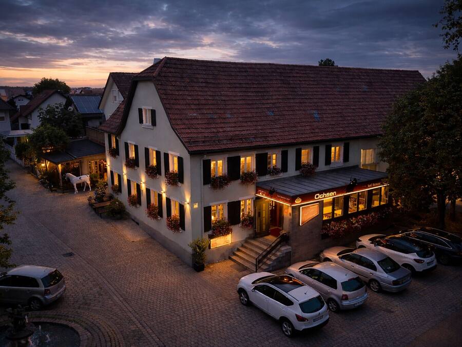 A two-story building with a red-tiled roof, a ground floor restaurant named "Ochsens," and a parking area with several cars.