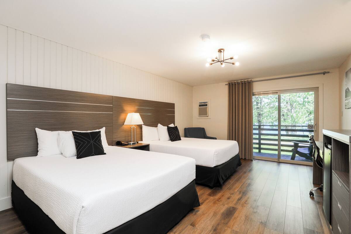 A hotel room with two beds, a desk, a chair, a lamp, and a view of the outdoors.