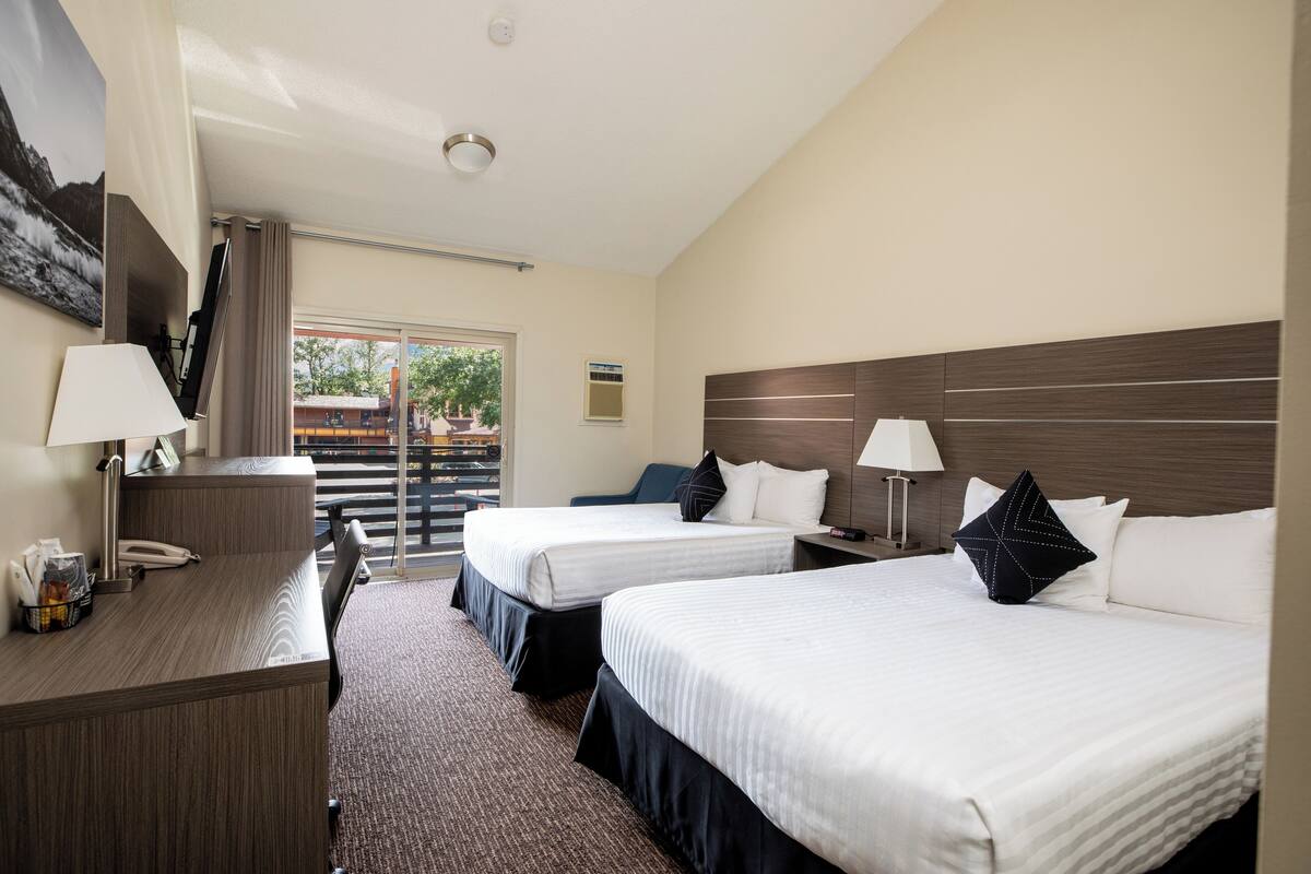 A hotel room with two beds, a desk, and a chair. There is a window with a view of the outside.