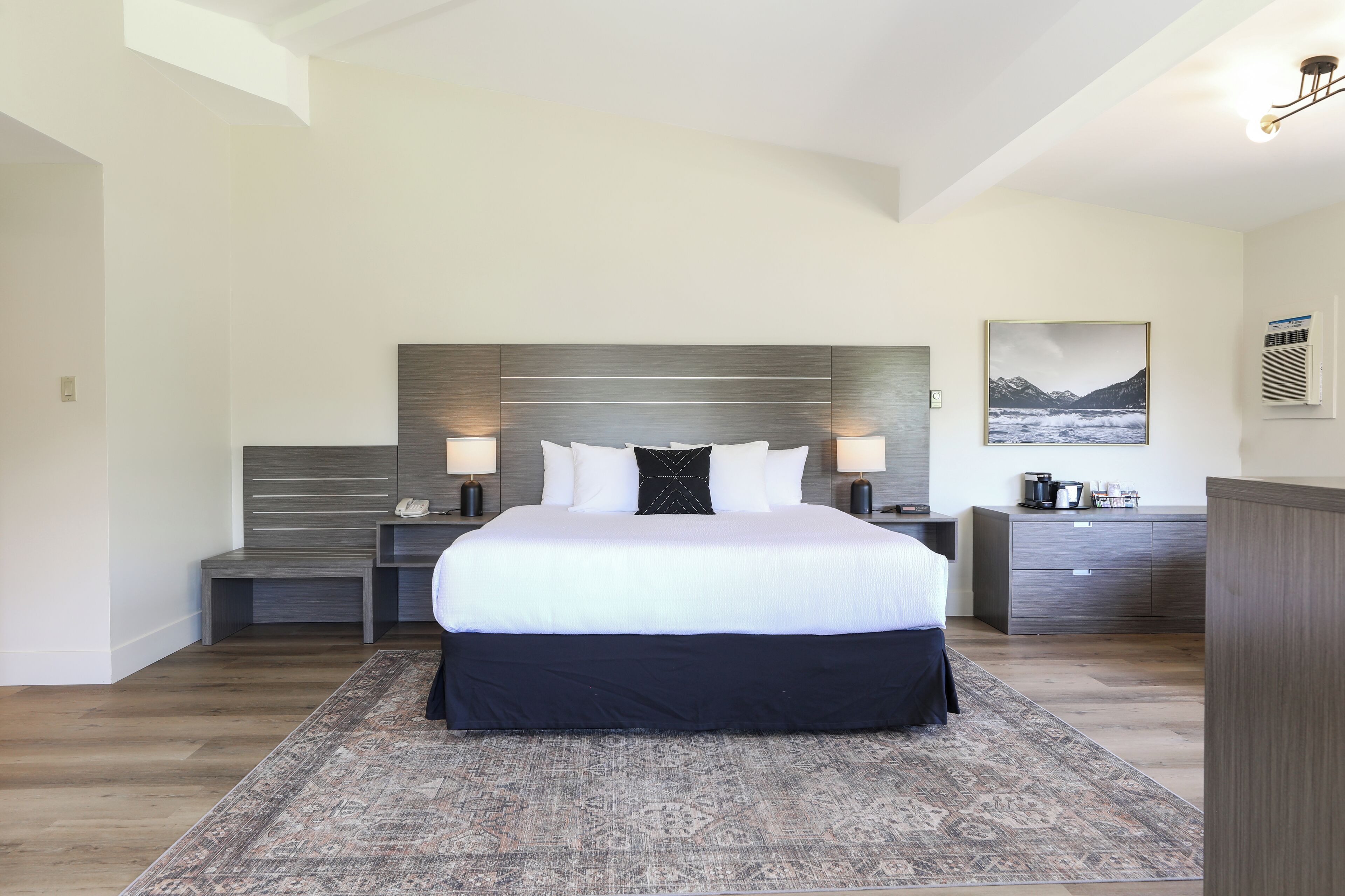 Ein modernes Hotelzimmer mit einem großen Bett, Nachttischen, einem Schminktisch und einem Wandbild von einer Berglandschaft.