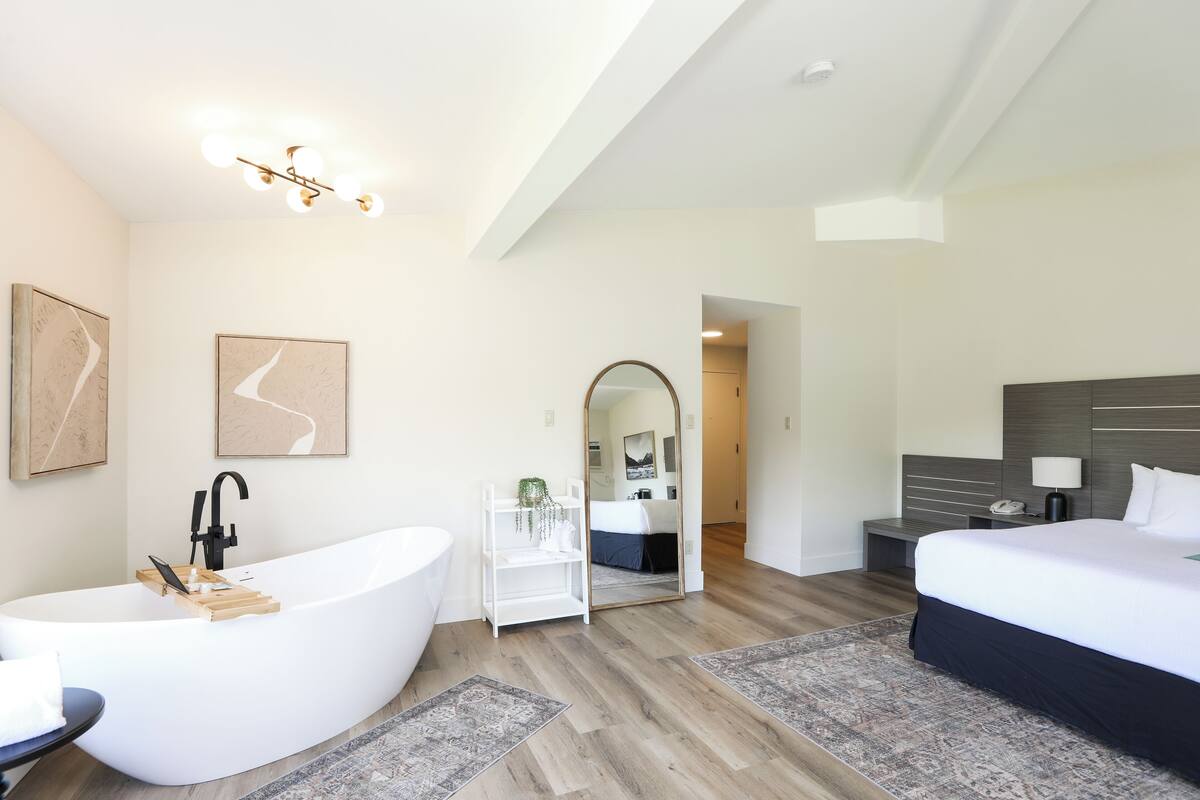 A modern hotel room with a freestanding bathtub, a large bed, a bedside table with a lamp, and a mirror.