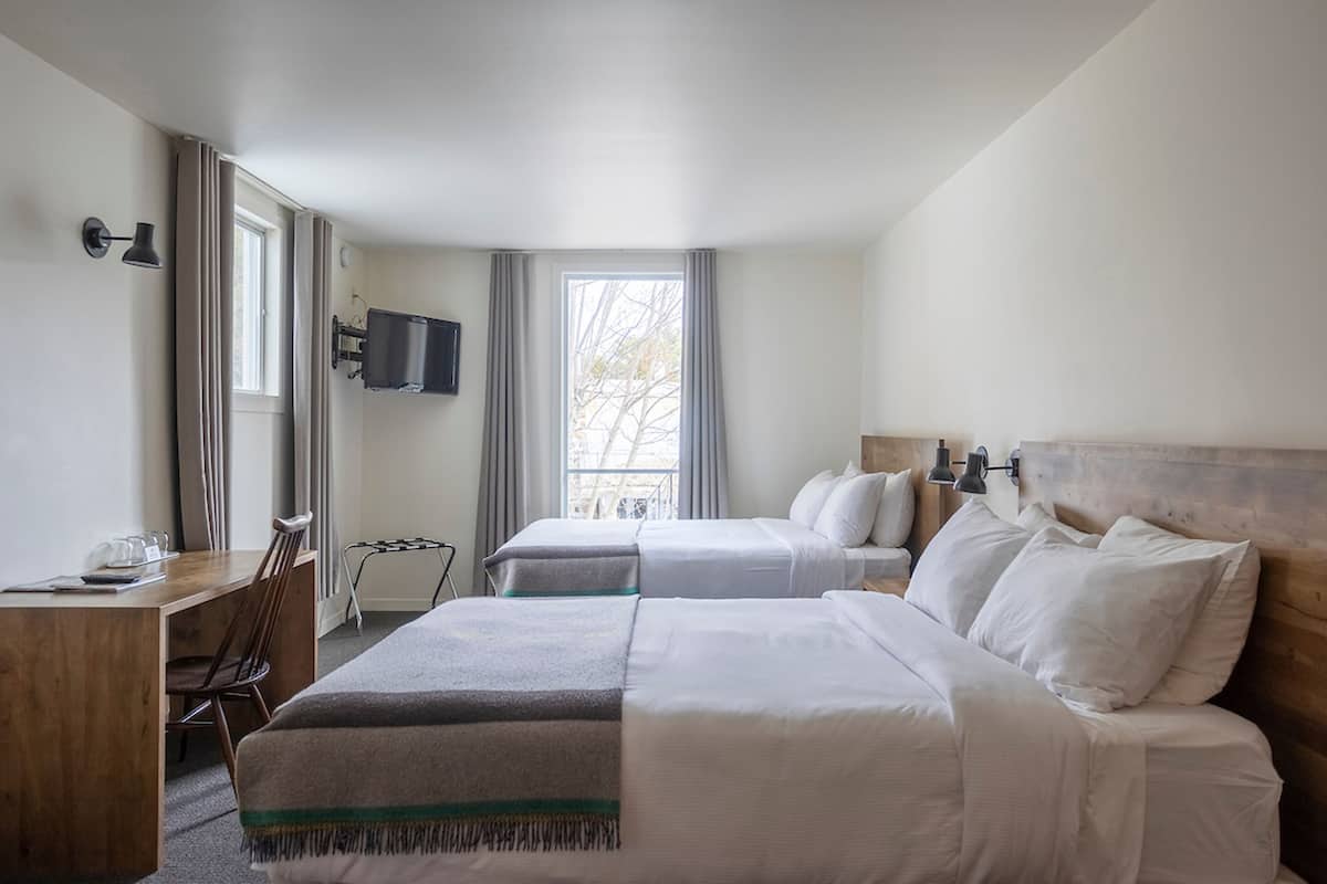 Habitación de hotel con dos camas, un escritorio, una silla, un televisor montado en la pared y un ventanal con cortinas.