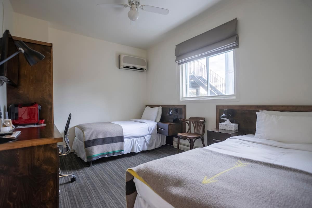 Habitación de hotel con dos camas, un escritorio, una silla, unidad de aire acondicionado y ventana con vista a un edificio.