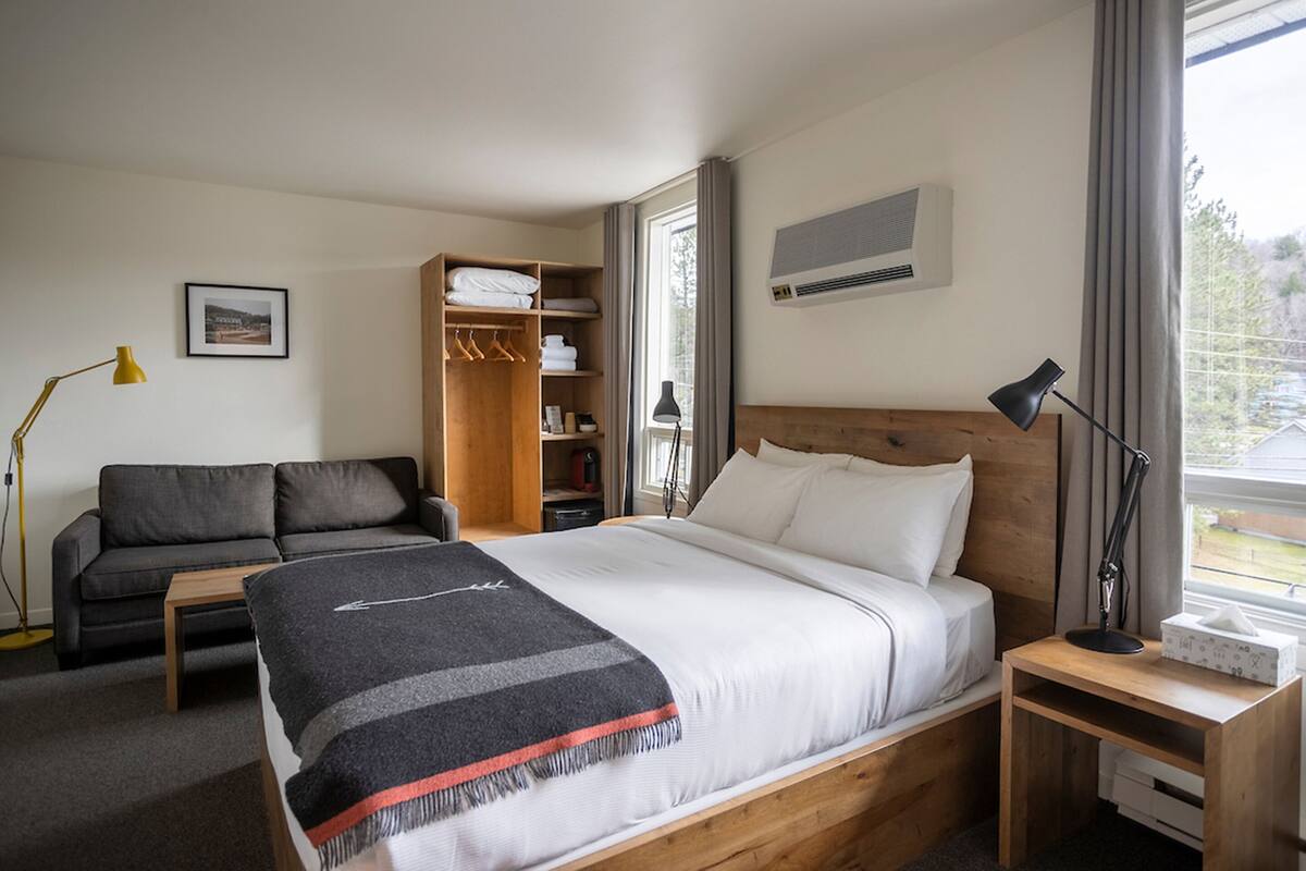 Habitación de hotel con cama, mesita de noche de madera, escritorio, silla, sofá, armario y ventana con cortinas.