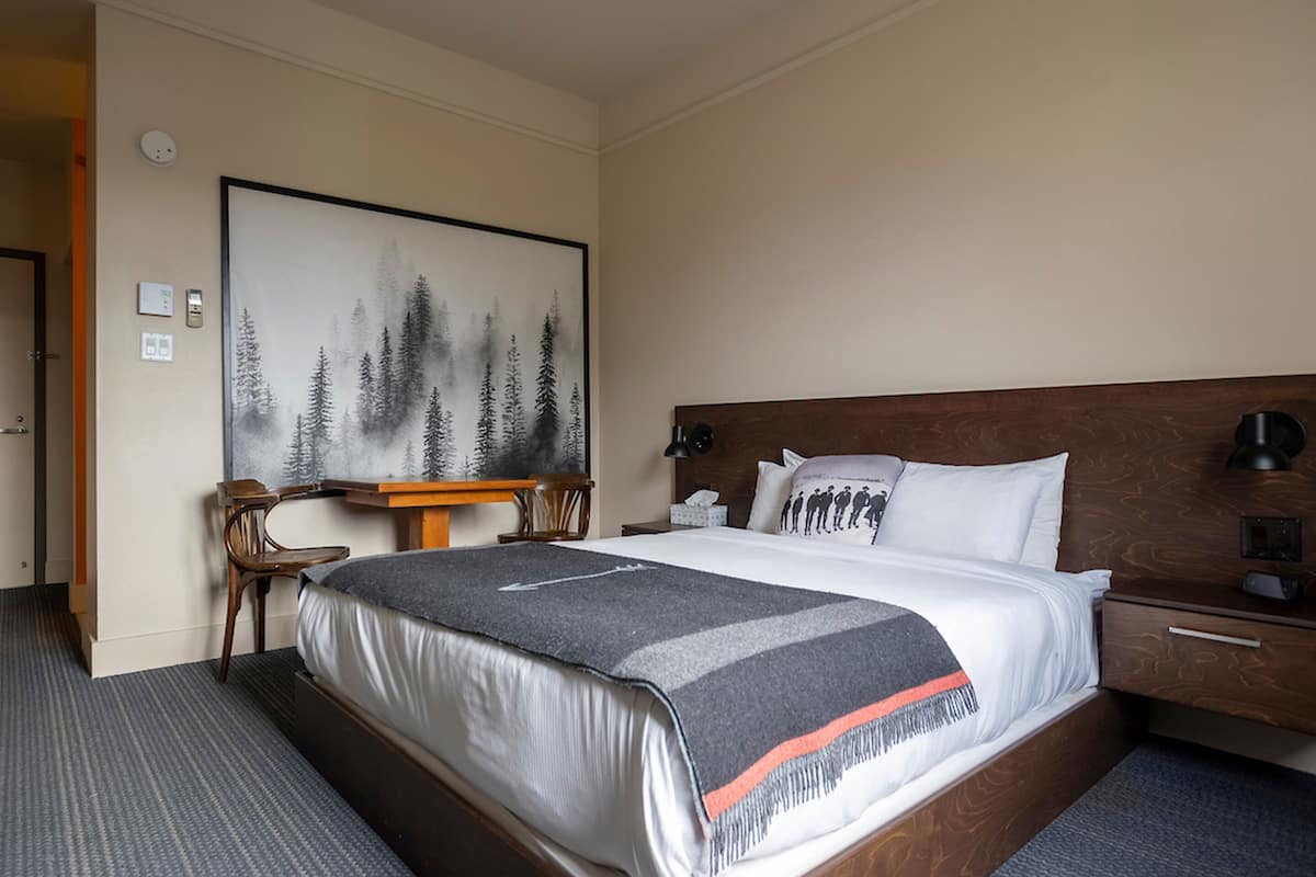 Habitación de hotel con una cama grande, un cabecero de madera, un escritorio con una silla, un cuadro en la pared y una mesita de noche con una lámpara.