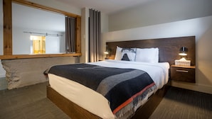 Ein Hotelzimmer mit einem großen Bett, einem hölzernen Kopfteil, Nachttischlampen, einem Beistelltisch und Blick auf den Flur durch ein Fenster.