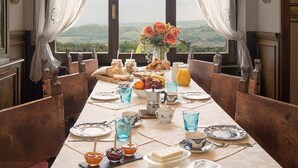 Comedor con una larga mesa dispuesta para una comida, ventanas grandes con cortinas y vistas al campo.
