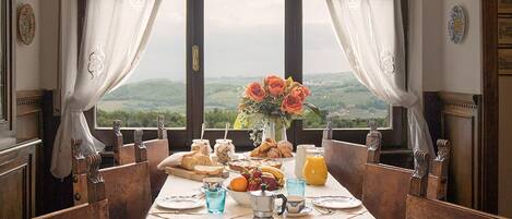 A dining room with a long table set for a meal, large windows with curtains, and a view of the countryside.
